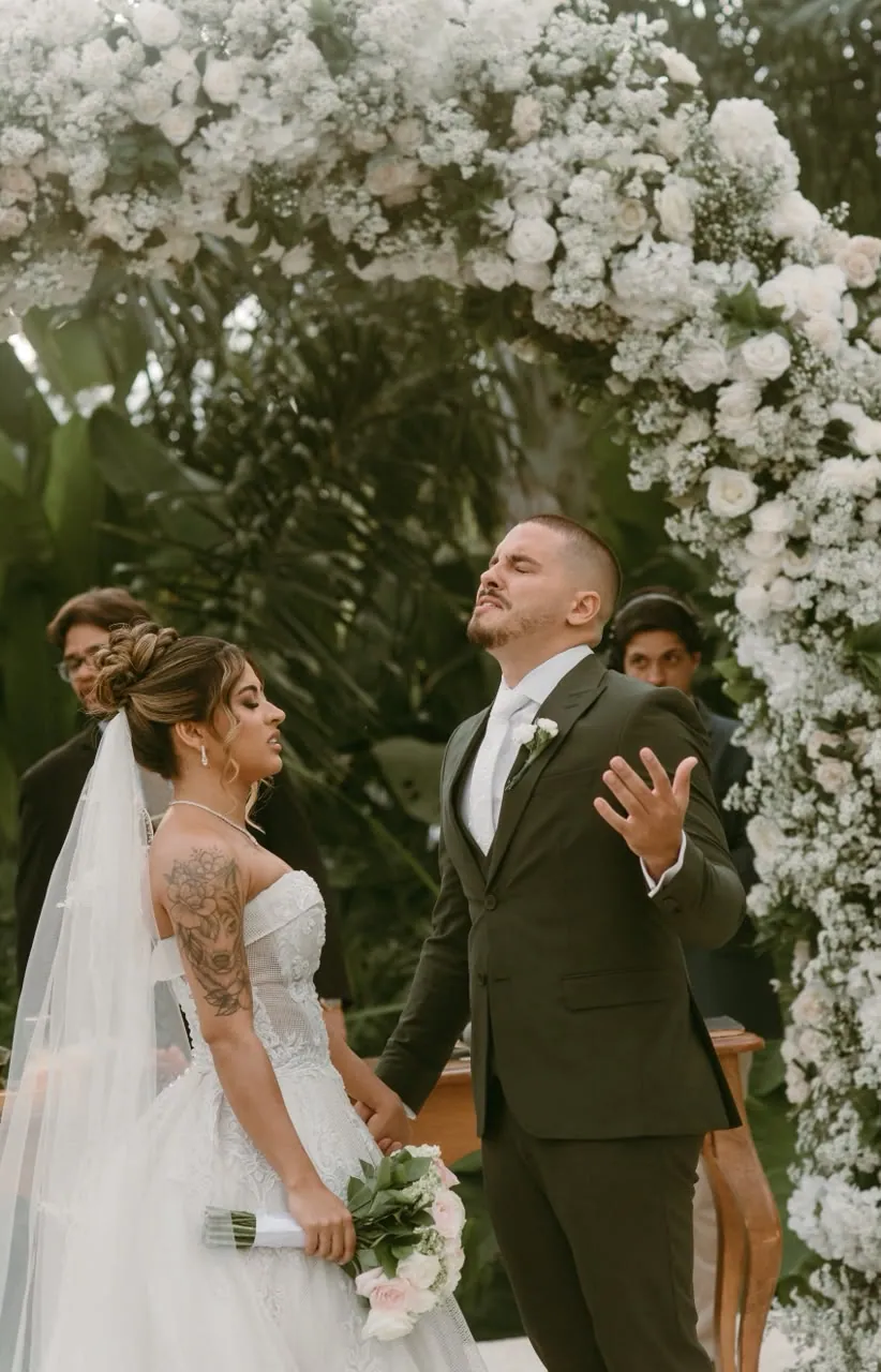 Joyce e Felipe durante a cerimônia sob arco de flores brancas em jardim tropical