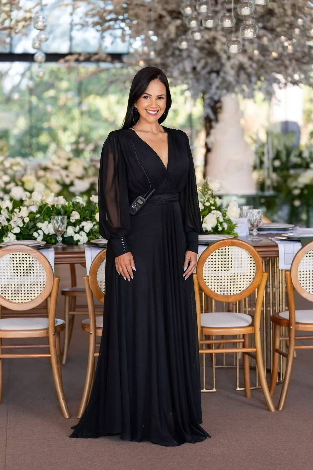 Cris Marques em vestido preto à frente da mesa dos noivos em casamento sob estrutura de vidro